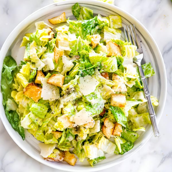 A close-up of a fresh Caesar salad with romaine lettuce, croutons, and Parmesan cheese on a white plate.